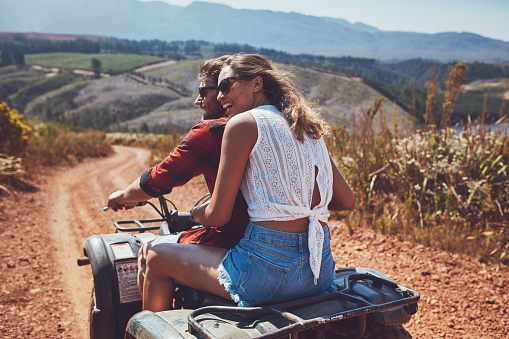 Rear view shot of young couple riding on a quad bike in countryside and looking away smiling. Woman sitting behind her boyfriend driving an quad on country road.