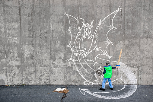 Little 5 year old boy fighting an imaginary dragon using a wooden sword and kitchen accessories as armor. Importance of reading and existence of non-visual media. Dragon chalk drawing on the concrete wall. Natural light.