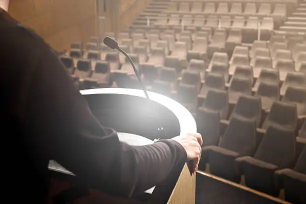 Close-up of unrecognizable man speaker on pupitre, testing its speech before conference, with microphone front empty seating of a common amphitheater.