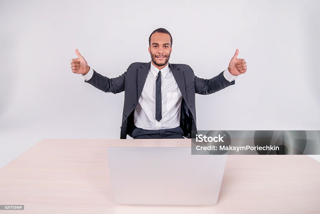 Serious businessman. Smiling African businessman sitting at a de Serious businessman. Smiling African businessman sitting at a desk on a laptop while businessman sitting at the table and showing thumb up over a laptop isolated on a gray background Adult Stock Photo