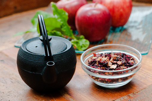 Red fruit infused tea in a glass ramekin, cast iron teapot & red apples on wooden table.