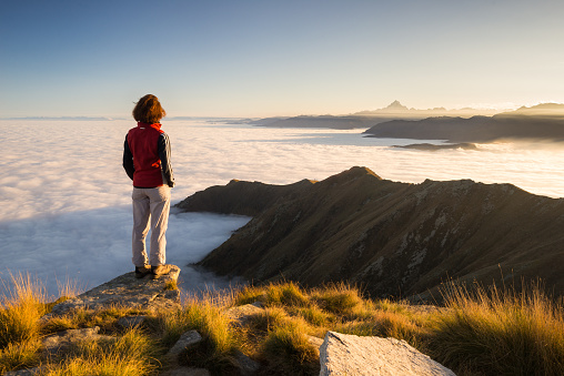 Female hiker reaching her goal at the mountain top and looking at majestic panoramic view of the italian western Alps with M. Viso peak in the background. Wide angle view at sunset.
