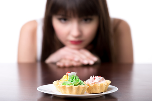 Young dieting woman sitting in front of plate with delicious cream tart cakes, looking at forbidden food with unhappy and hungry expression, studio, white background, isolated, close-up