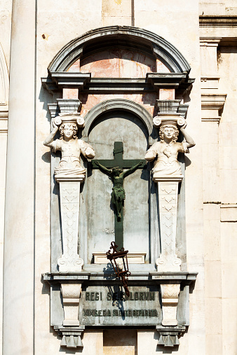 Christian sculptures at facade of old building in Città Alta of Bergamo. In center is Jesus at cross framed by two columns with women. Latin text below. Facade is close to old library.