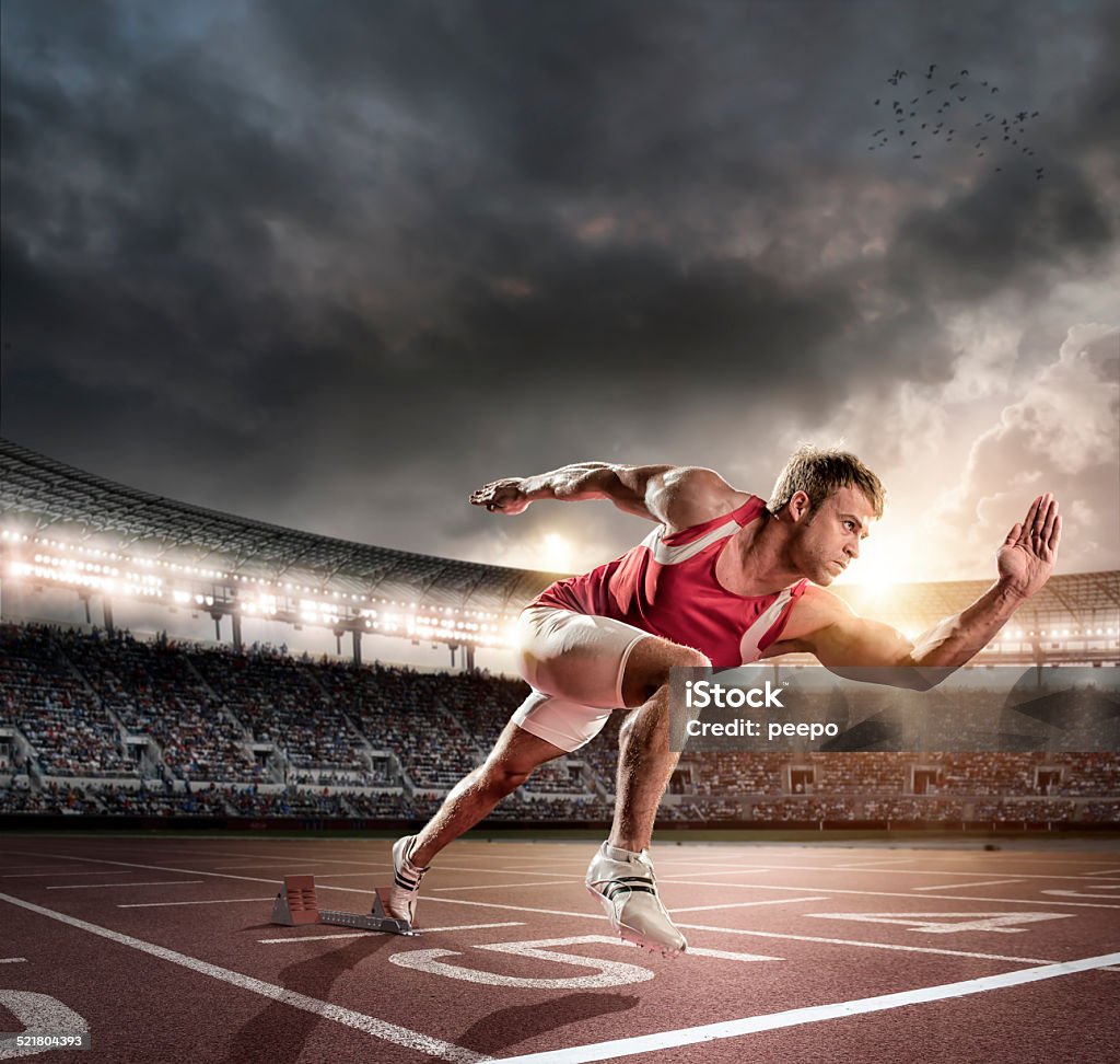 Sprinter Bursting from Starting Blocks on Track A mid action image of professional male athlete sprinting out of starting blocks on numbered outdoor running track. Action takes place in a generic floodlit outdoor athletics arena full of spectators under a stormy evening sky at sunset. Sprinter is wearing generic unbranded athletics strip. Composite image with intentional lensflare. Running Stock Photo