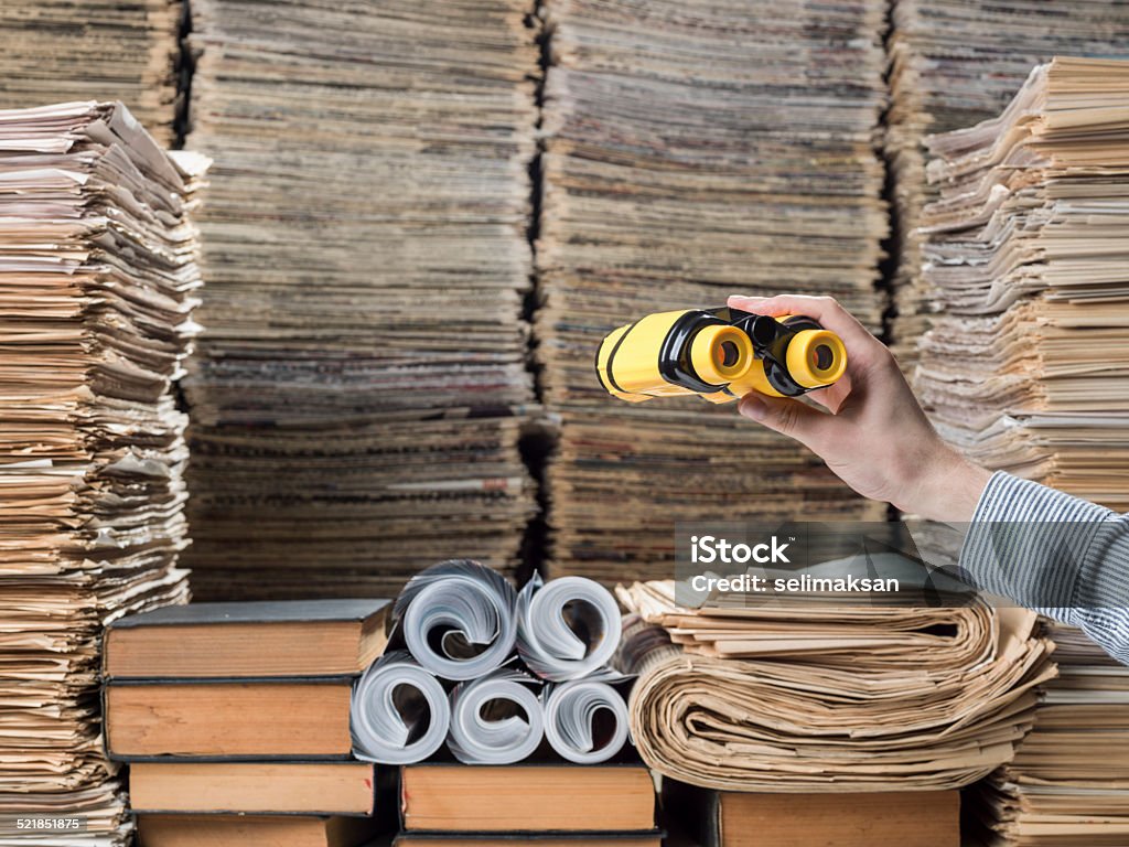 Binoculars in human hand and searching messy library Yellow binoculars in human hand and searching messy library.Human hand is seen on the right side of frame.Newspapers,magazines,books and documents are stacked on shelfs.Some magazines are rolled.The photo was shot with a medium format DSLR camera Haseelblad H4D in studio. Archives Stock Photo