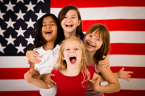 Color photo of four happy, patriotic, young girls laughing together in front of a large American flag. Differing ethnicity portraying unity, friendship, acceptance and peace.