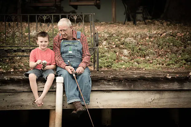 Color image of a senior man sitting on an old, wooden dock while fishing with his young great grandson.