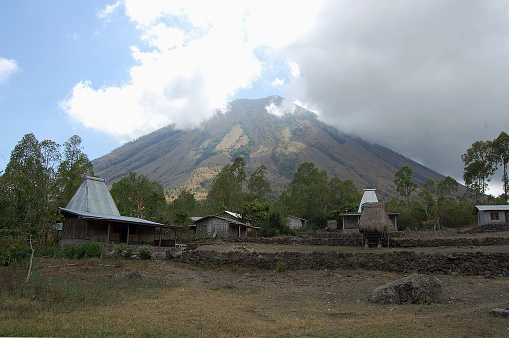 Inerie volcano, semi covered by clouds surrounding tradtional wooden houses. It's a 2245 meters high volcano with an impressive pyramid shape and is located close to the town of Bajawa. Flores island