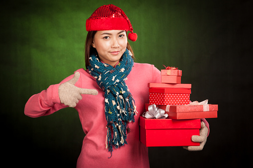 Asian girl with red christmas hat point to gift boxes on dark green background