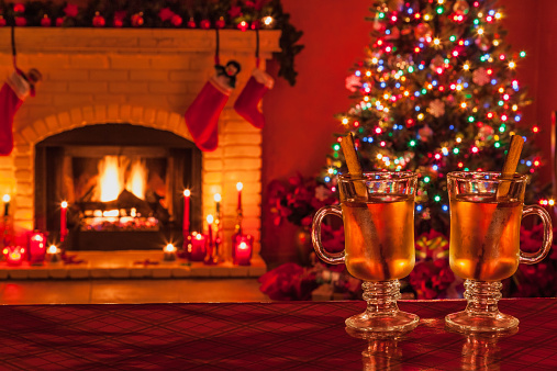Two glass mugs of hot apple cider with cinnamon sticks sit on table in front of a Christmas scene of Christmas tree, decorations, and fireplace with stockings, at night with a warm cozy lighting.