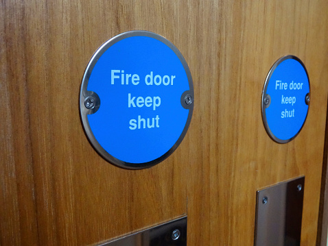 Photo showing a circular blue sign with white writing saying 'Fire Door Keep Shut '.  This safety sign has been screwed onto a wooden fire door.