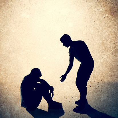 A silhouette of a man reaching out to help assist another person who is sitting on the ground with his head looking toward the ground in sadness or depression.  Meant to depict aid, assistance, charity, relief work, and care for the needy, homeless, and troubled. Square crop with copy space.  Textured background.