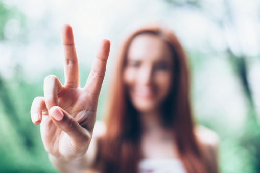 Redheaded young woman making victory sign with her hand and smiling. Blurred background.