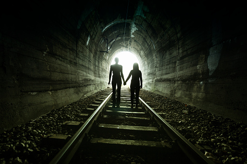 Couple walking hand in hand along the track through a railway tunnel towards the bright light at the other end, they appear as silhouettes against the light