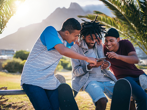 Multi-Ethnic group of skater friends hanging out, sitting on rail at seaside with skateboards resting against legs, while looking at a smartphone, pointing and laughing during sunset