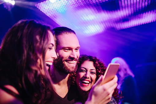 Happy young adult friends laugh looking at pictures on a smarphone during a party in a night club