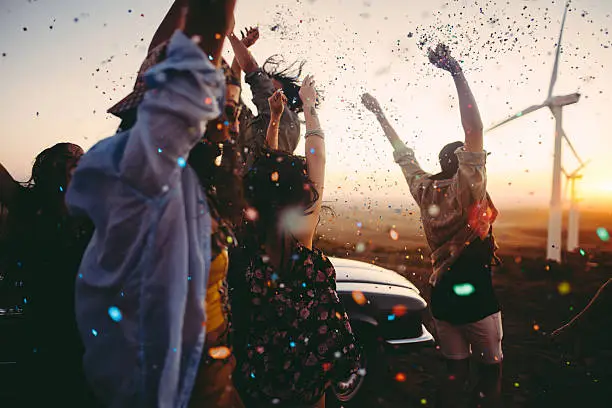 Group of multi-ethnic teens dressed in boho style partying outdoors throwing colourful confetti in a wind farm at sunset during their american carefree road trip