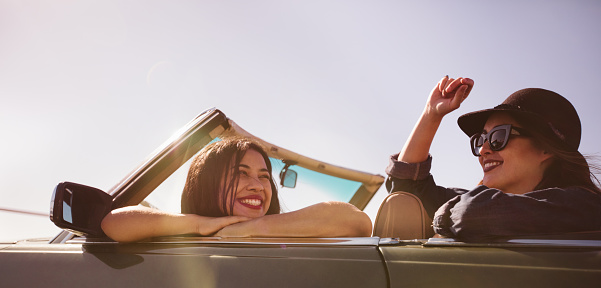 Two cheerful teenager friends smiling and enjoying a summer road trip on a vintage convertible car