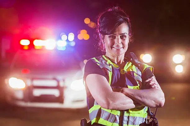 A female police officer standing in the street at night, arms folded, smiling and looking at the camera. Her patrol car is in the background with the emergency lights illuminated.
