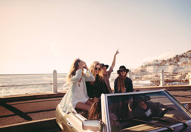 Friends happy to reach the sea on their road trip Group of multi-ethnical teenager friends smiling and celebrating in a convertible parked close to the beach hipster car stock pictures, royalty-free photos & images