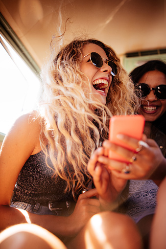 Boho or hipster fashion woman laughing out loud looking at her smart phone with mixed race girl friend in vintage van during a road trip.