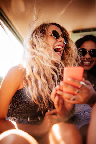 Hipster girl looking at smart phone with road trip friend Boho or hipster fashion woman laughing out loud looking at her smart phone with mixed race girl friend in vintage van during a road trip. hipster car stock pictures, royalty-free photos & images