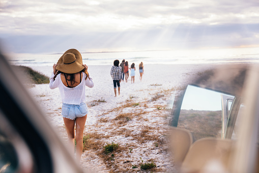 A Hipster multi-ethnic group of friends walking barefoot and happy from parked retro van on to sandy beach while on ocean road trip