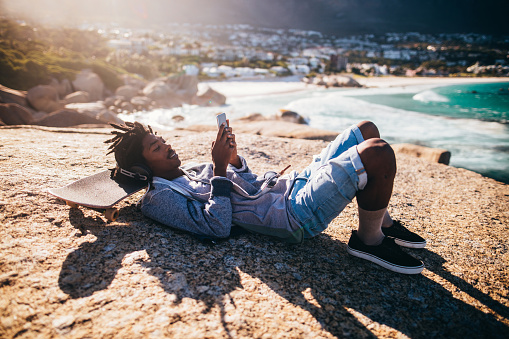 African American skater lying down, listening to music from his smartphone with headphones and resting his head on his skateboard on the rocks of a seaside during sunset
