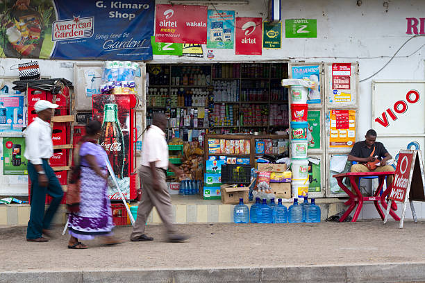 Mwanza, Tanzania: Pedestrians Walk By Small Shop Mwanza, Tanzania - May 10, 2012: Pedestrians in central Mwanza walk by small shop tended by a young man sitting out front. mwanza city tanzania stock pictures, royalty-free photos & images