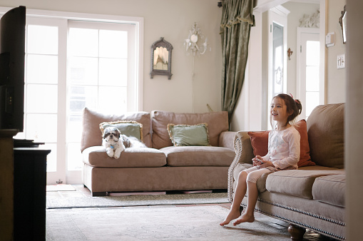 A pretty little girl is holding and using an Apple TV remote while watching TV.  She appears happy and entertained by what she's watching. She's 6 years old and Eurasian. Her dog is sitting on the couch in the background. The home's interior appears stylish and includes old-world/vintage home decor.