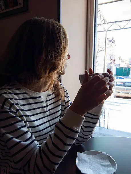 Young woman with coffee cup in hand sitting near the window at a table in cafe