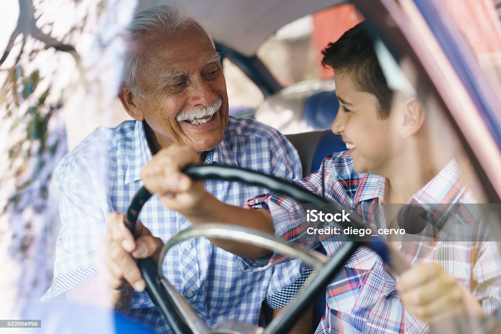 Old Man Grandpa Gives Driving Class To Grandson Family and Generation gap. Old grandpa spending time with his grandson and teaching him to drive. The boy holds the volante of a vintage car from the 60s. They both smile happy looking each other. Grandfather Stock Photo
