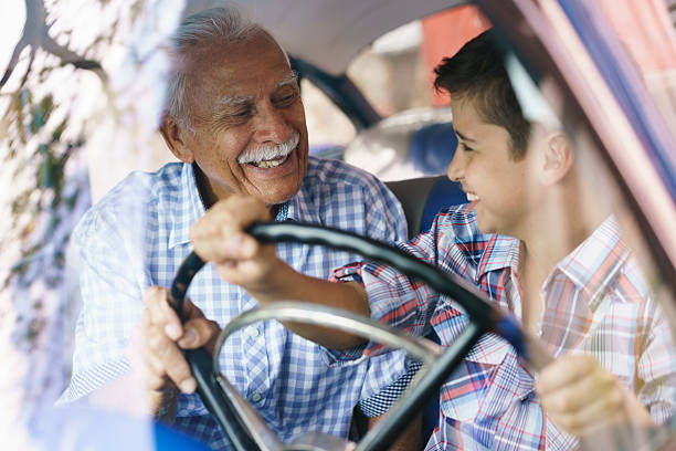 Old Man Grandpa Gives Driving Class To Grandson Family and Generation gap. Old grandpa spending time with his grandson and teaching him to drive. The boy holds the volante of a vintage car from the 60s. They both smile happy looking each other. grandson photos stock pictures, royalty-free photos & images
