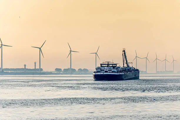 Rear view on a barge sailing in harbour at sunrise, wind turbines in a row along the water's edge in the background in Rotterdam The Netherlands