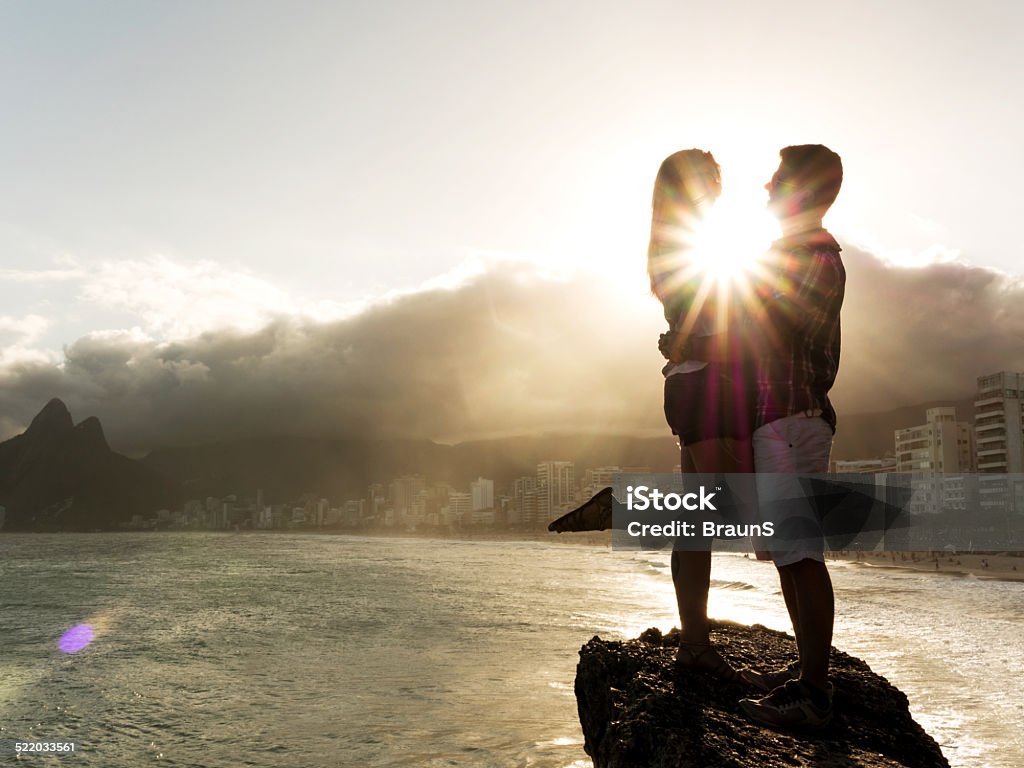 Romantic couple on a rock at sunset. Couple standing embraced on a rock near the beach and looking at each other. Copy space. Adult Stock Photo