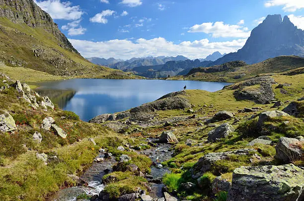 A mountain stream runs to the alpine lake Gentau. There are tourists and the recognizable summit Pic du Midi d'Ossau in the background.