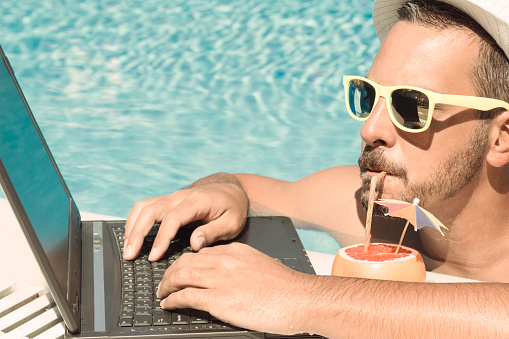Man working on laptop at the poolside while drinking fresh grapefruit juice by using a drinking straw  