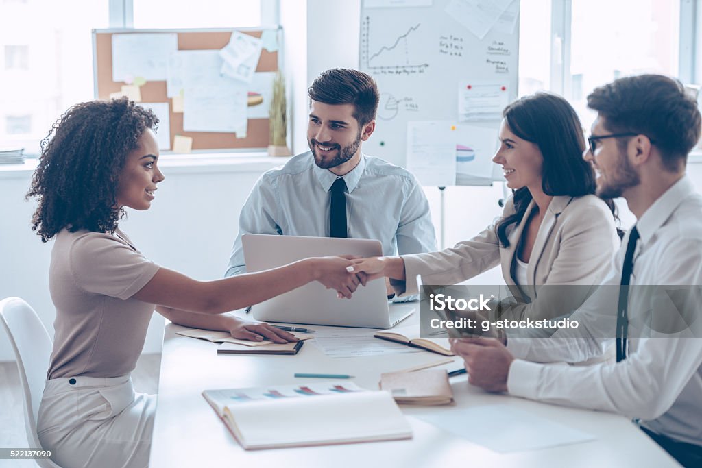 You are hired! Two beautiful women shaking hands with smile while sitting at the office table with their coworkers Business Finance and Industry Stock Photo