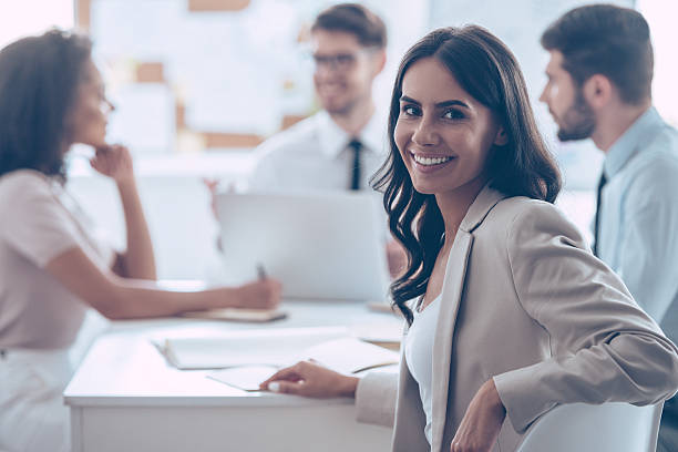 Feeling satisfied with her team. Beautiful cheerful woman looking at camera with smile while sitting at the office table with her coworkers stubble male african ethnicity facial hair stock pictures, royalty-free photos & images