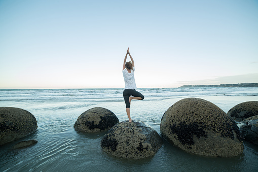 Young woman exercises yoga on a boulder at Moeraki boulders. She is holding the famous tree pose, shot at dusk.