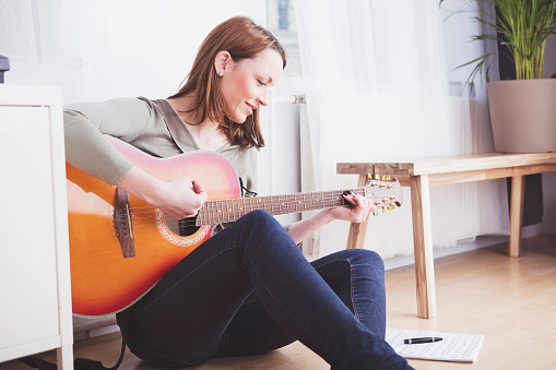 Pretty casual dressed woman with long brown hair sitting on the floor of the living room playing some records on guitar