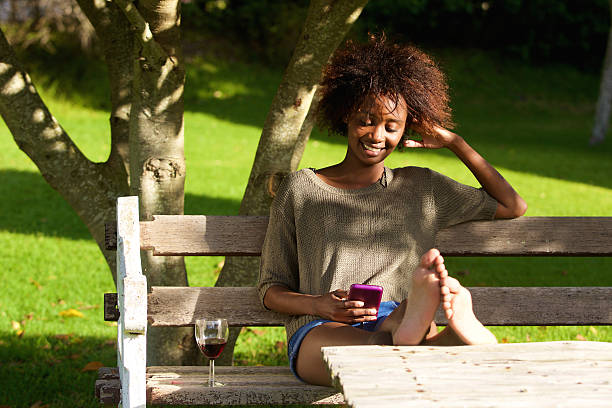 mulher sorridente preto sentado no parque com telefone móvel - using phone garden bench imagens e fotografias de stock