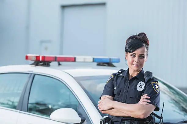 A policewoman standing next to her patrol car, smiling and looking at the camera with her arms crossed. She is in the standard navy blue policeuniform.