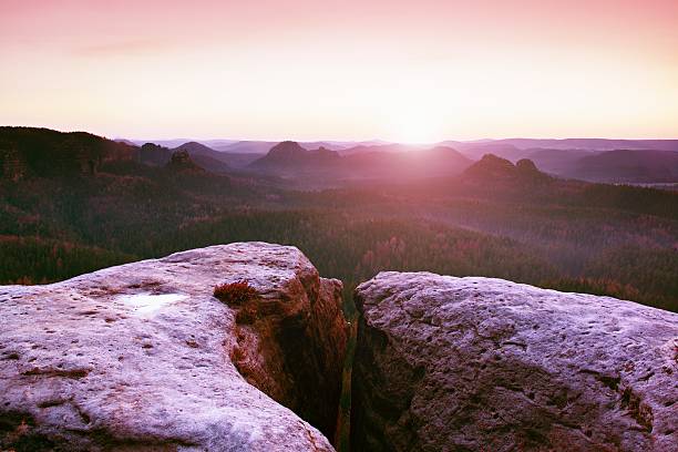 Morning view over sandstone cliff into forest valley, daybreak Sun at horizon. Hills increased from foggy background