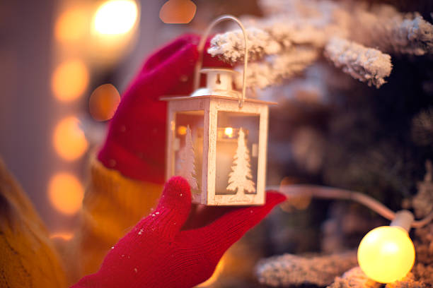 Close up of woman's hands holding lantern, standing by christmas tree. Wearing knitted sweater and gloves. Tree is decorated with festive string lights. Evening or night with beautiful yellow lights lightning the scenes. FOCUS on hands.