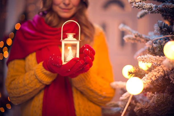 Close up of woman's hands holding lantern, standing by christmas tree. Wearing knitted sweater and gloves. Tree is decorated with festive string lights. Evening or night with beautiful yellow lights lightning the scenes. FOCUS on hands.
