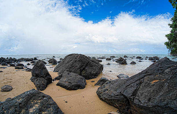 Image of lava rock and soft sand beach at Anahola Beach Park on the northeast side of Kauai, Hawaii.  This beach is well protected and local families will bring their children for a day at the beach.  People will sit in the shade and have a picnic or a barbque beside the surf gently breaking on the sandy shore.  Ironwood trees provide shade on the east side of the beach and locals will fish from the black lava rocks on that side of the beach.  A peaceful and tranquil location to spend the day or have a Hawaiin vacation.