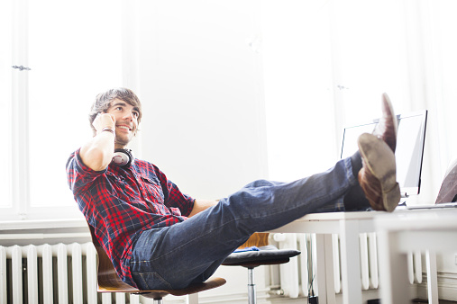 Relaxed young man talking on mobile phone at work with feet on desk