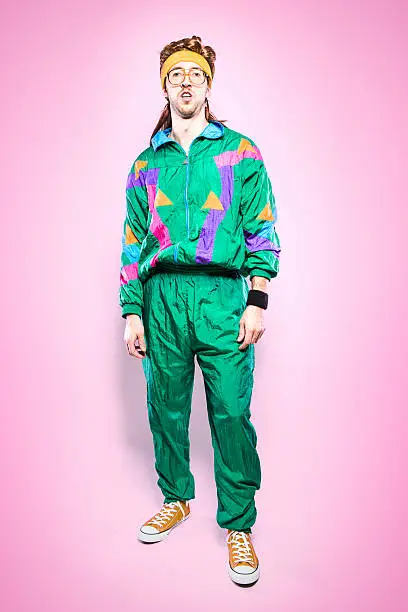 A cool, funky young adult in late 1980's / early 1990's fashion style, with mullet, fluorescent colored track suit, nerdy glasses, and sweat band.  Vibrant pink background. Vertical portrait.